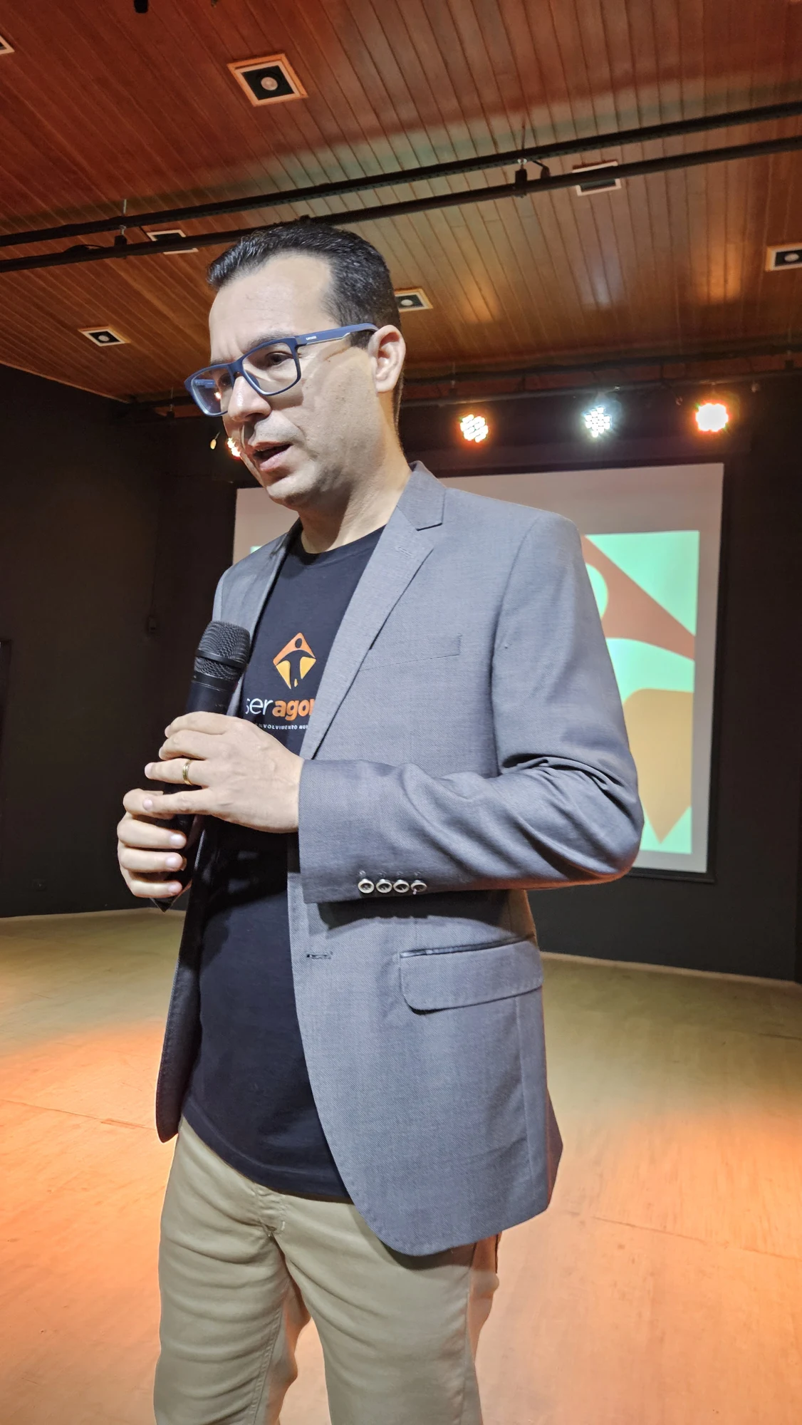 Gustavo Isaias fala ao microfone em um palco durante um evento